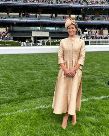 Woman in a beige dress and hat stands on grass in front of a stadium filled with people at an outdoor event on a sunny day.