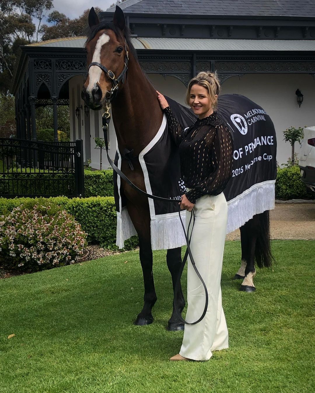A brown horse stands on a grassy lawn wearing a blanket, next to a fair-skinned woman in cream trousers and a black top.