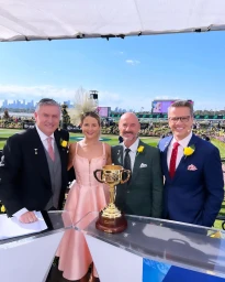 Four people standing behind a podium displaying a gold trophy at a horse racing event under a white canopy on a sunny day.