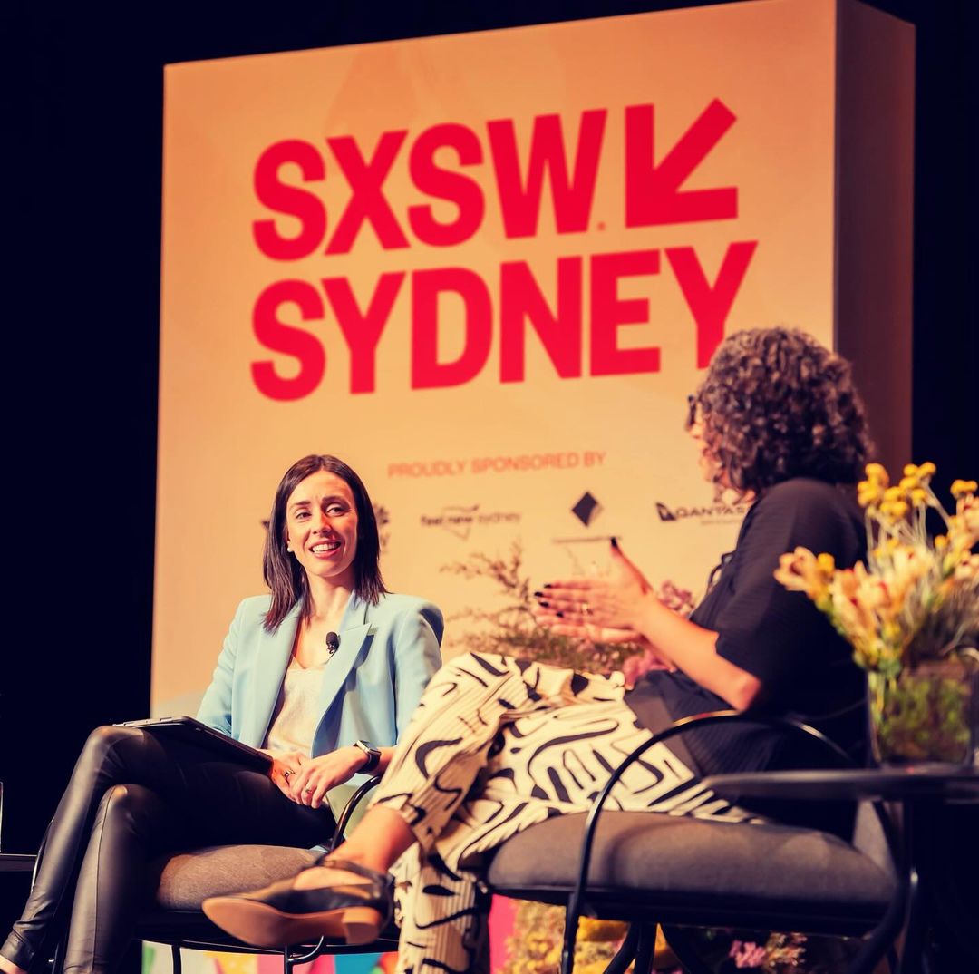 Two women are seated on chairs on stage at SXSW Sydney. One is wearing a blue blazer. The background features the SXSW logo.