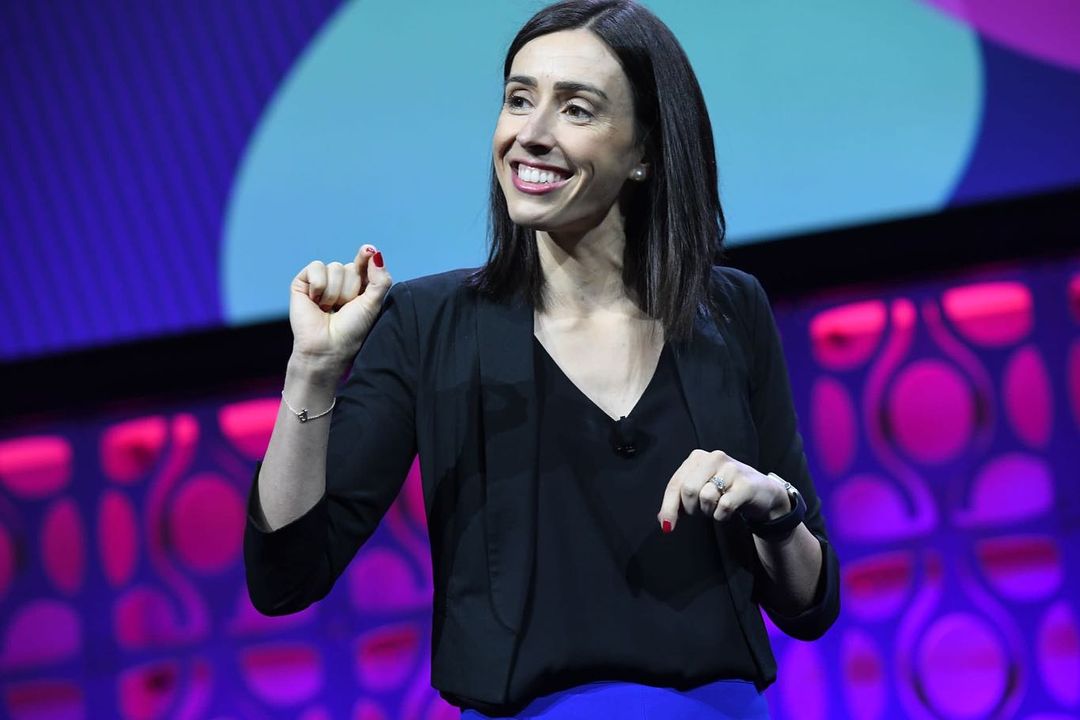 Smiling woman gestures on stage at an event. She wears a black jacket and blue skirt, with a patterned backdrop in pink and blue.