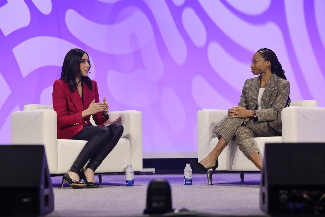 Two women sit on white armchairs on stage at a conference. One woman wears a red blazer, the other a plaid suit.