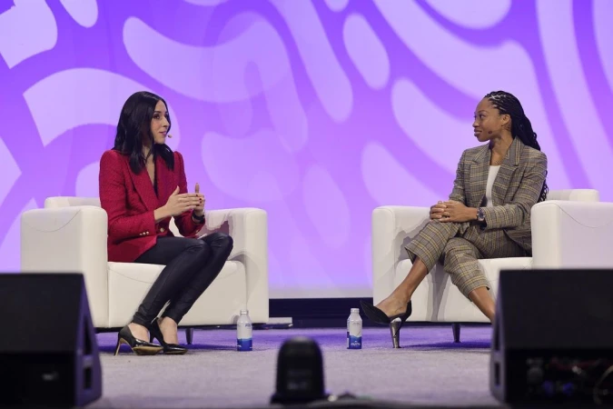 Two women sit on white armchairs on stage at a conference. One woman wears a red blazer, the other a plaid suit.