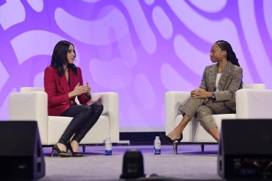 Two women seated in white armchairs on stage during a panel discussion, with a purple patterned backdrop.