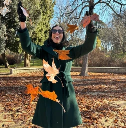 Woman in dark green belted coat throwing autumn leaves in the air outdoors in a park.