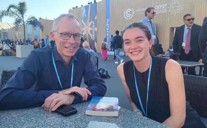 Two smiling people pose at an outdoor climate conference, with a book on the table and conference signage in the background.