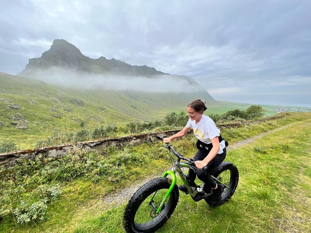 A woman rides a fat tire bike on a grassy trail with a mountain partially obscured by fog in the background on a cloudy day.