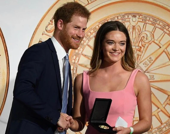 Two people shaking hands. A fair-skinned man in a suit and a woman in a pink dress hold a cased medal indoors on light backdrop.