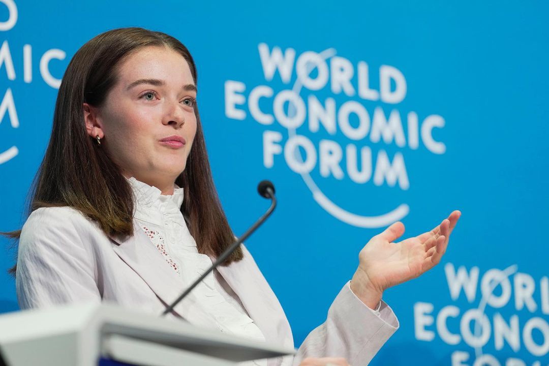 Brunette woman in blazer presents at World Economic Forum, with a microphone in front of a blue branded backdrop.