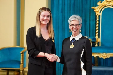 Two women shaking hands indoors against a teal curtain background, featuring content creator Jade Hameister in a black jacket.