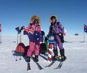 Two polar explorers in winter gear on skis standing next to an Australian flag and a marker in a snowy landscape under a clear blue sky