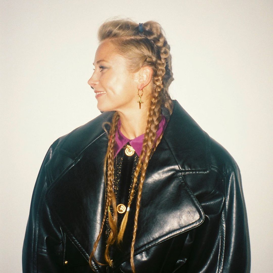 Profile of a fair-skinned woman with blonde braided hair wearing a black leather trench coat with gold buttons against white backdrop.