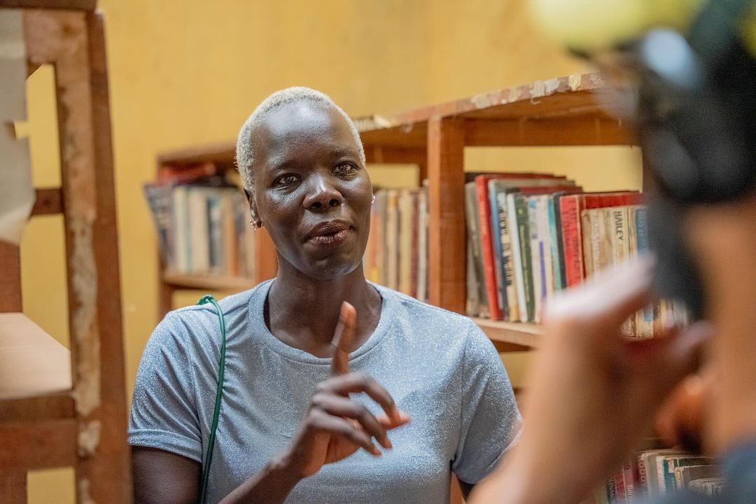 An African woman with platinum blonde hair stands indoors near wooden shelves holding books, while another person appears out of focus.