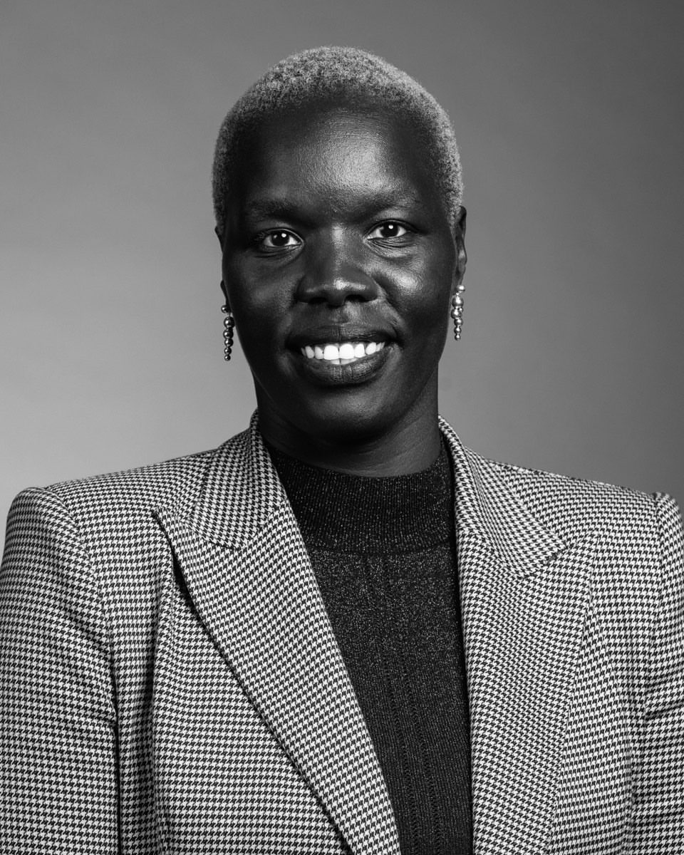 Black and white portrait of an adult woman with short blonde hair smiling in a photo studio, wearing a patterned blazer and a dark shirt.