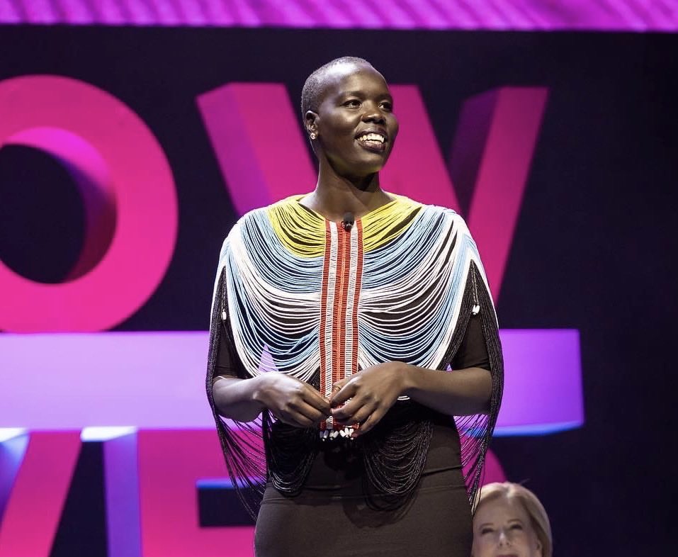 Woman standing on stage wearing a beaded top, looking toward the right. Stage background with large pink letters.