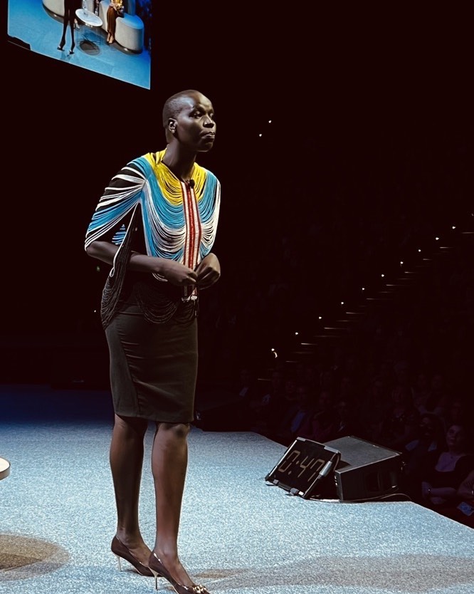 Adult female stands on stage in dark dress and colorful fringed cape. Event platform with audience seating in blurred background.