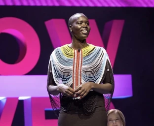 Nyadol Nyuon smiling on stage wearing a beaded fringe collar necklace over a dark dress against a pink and white backdrop