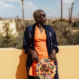 Nyadol Nyuon wearing an orange dress and dark blue robe holding a patterned fan outdoors