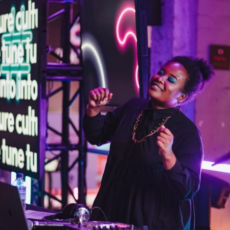 Enthusiastic Black female DJ with bright blue eyeshadow dances behind sound mixing equipment against LED sign backdrop in nightclub.