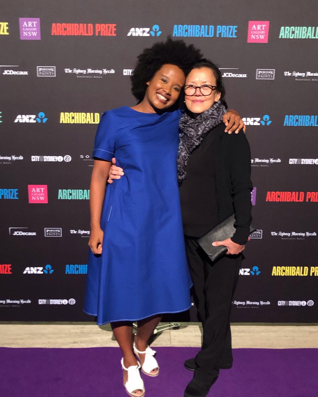 Two women pose at the Archibald Prize event, one in a blue dress and white heels, the other in black attire, against branded backdrop.