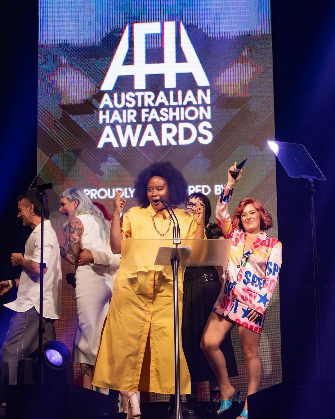 Presenter at Australian Hair Fashion Awards stands on stage, smiling and gesturing next to the awards logo, with attendees on either side.