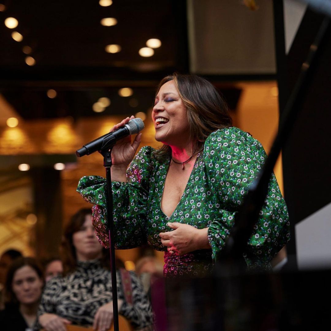 A woman with dark hair sings into a microphone while wearing a floral green dress in an indoor venue with an audience.