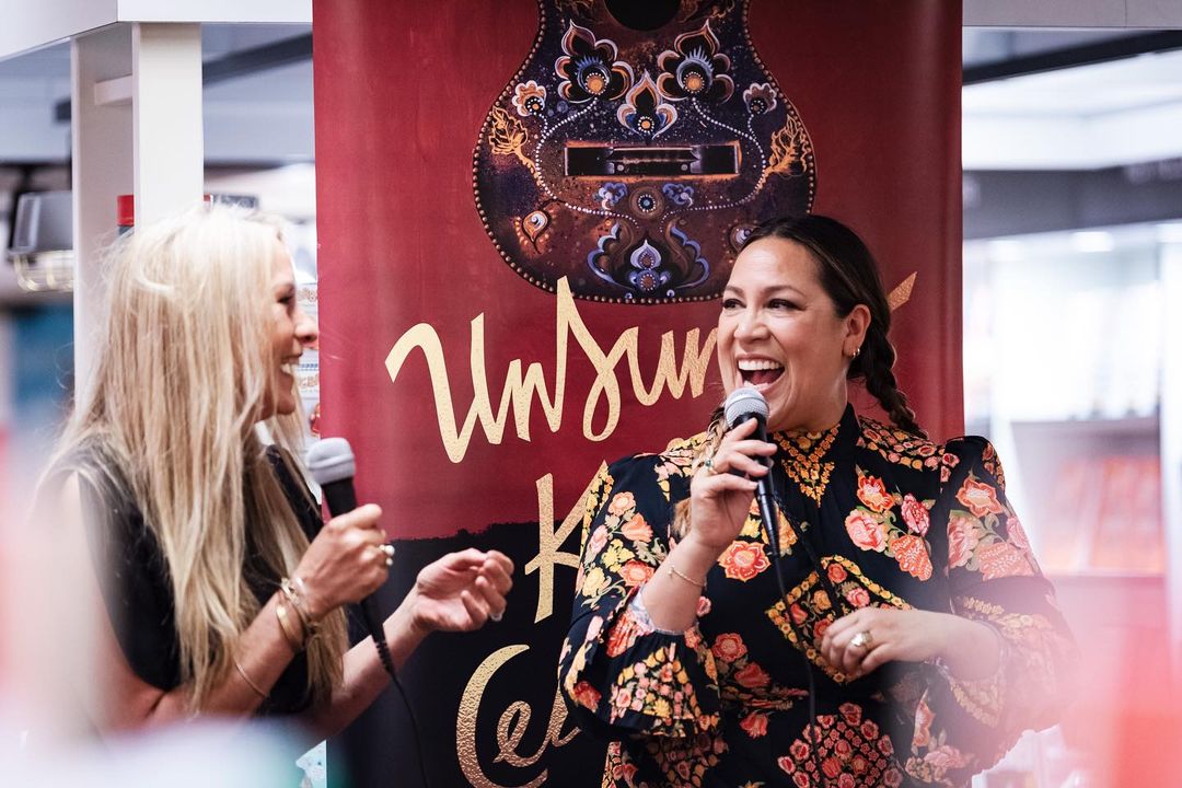Two women joyfully speak into microphones at an event; one in a floral dress and the other with long blonde hair.