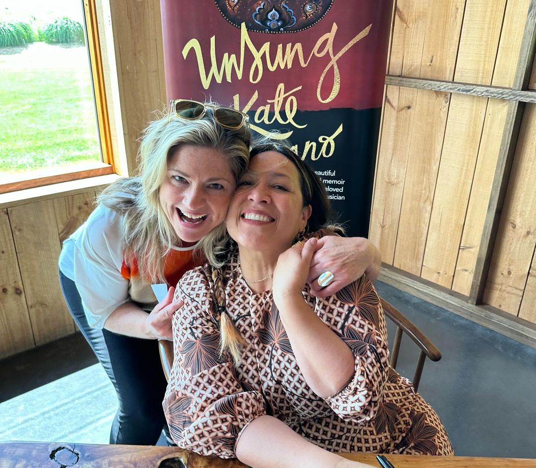 Two smiling women pose for a photo indoors, with one seated and the other standing with an arm around her. A book cover stands in the background.