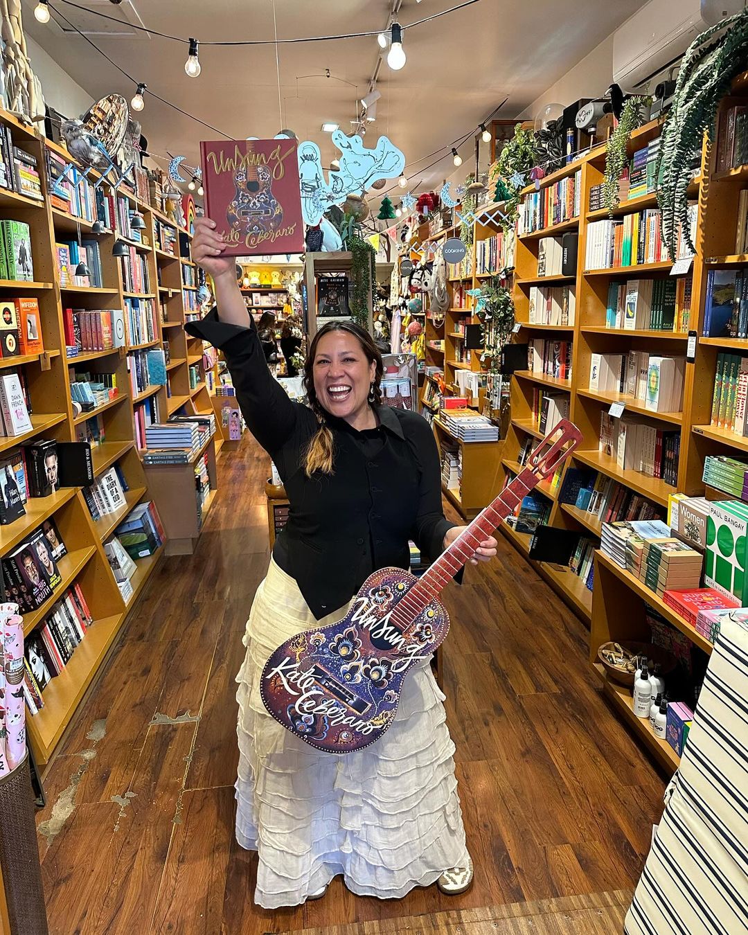 Enthusiastic woman in a bookshop raising her book and holding a decorative guitar, creating a joyous scene among rows of books.