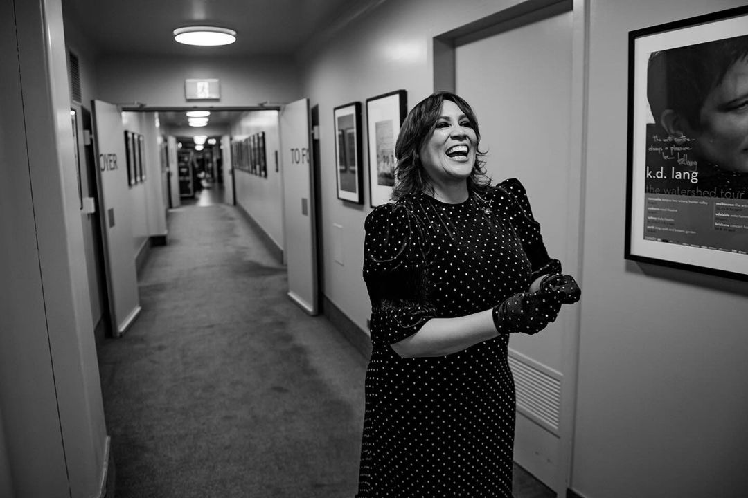 Black and white shot of a smiling adult woman in a polka dot dress in a hallway with posters and doorways in the background.