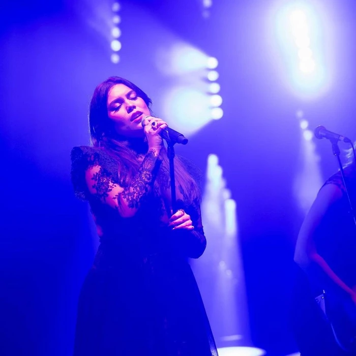 Kate Ceberano singing into a microphone on stage under dramatic blue lighting with soft bokeh background spots.