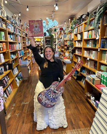 Content creator Kate Ceberano holding a book and guitar in a bookstore with wooden bookshelves.
