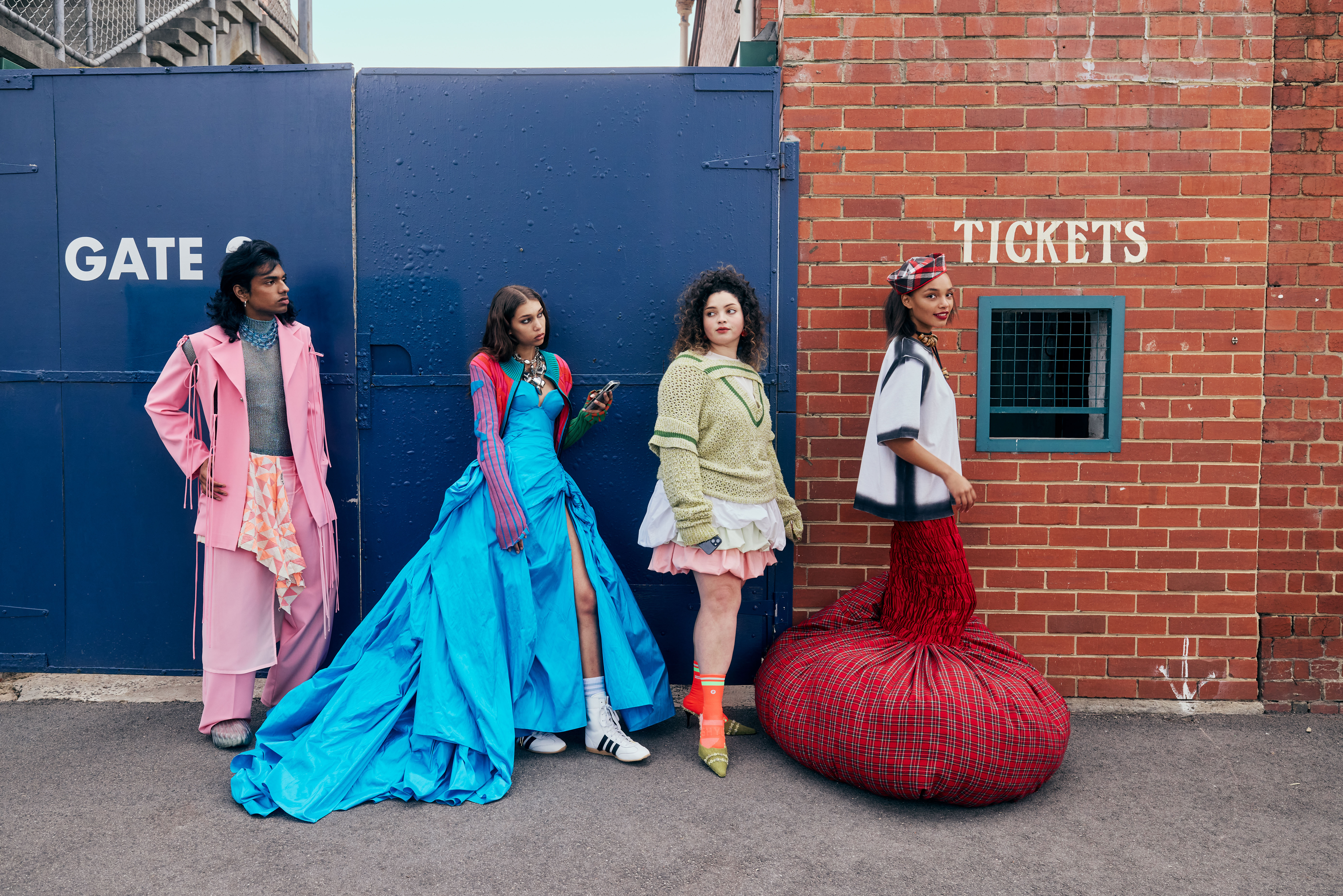 Four diverse models showcase fashion in front of a blue gate and brick wall with a ticket window, creating an eye-catching editorial shot.