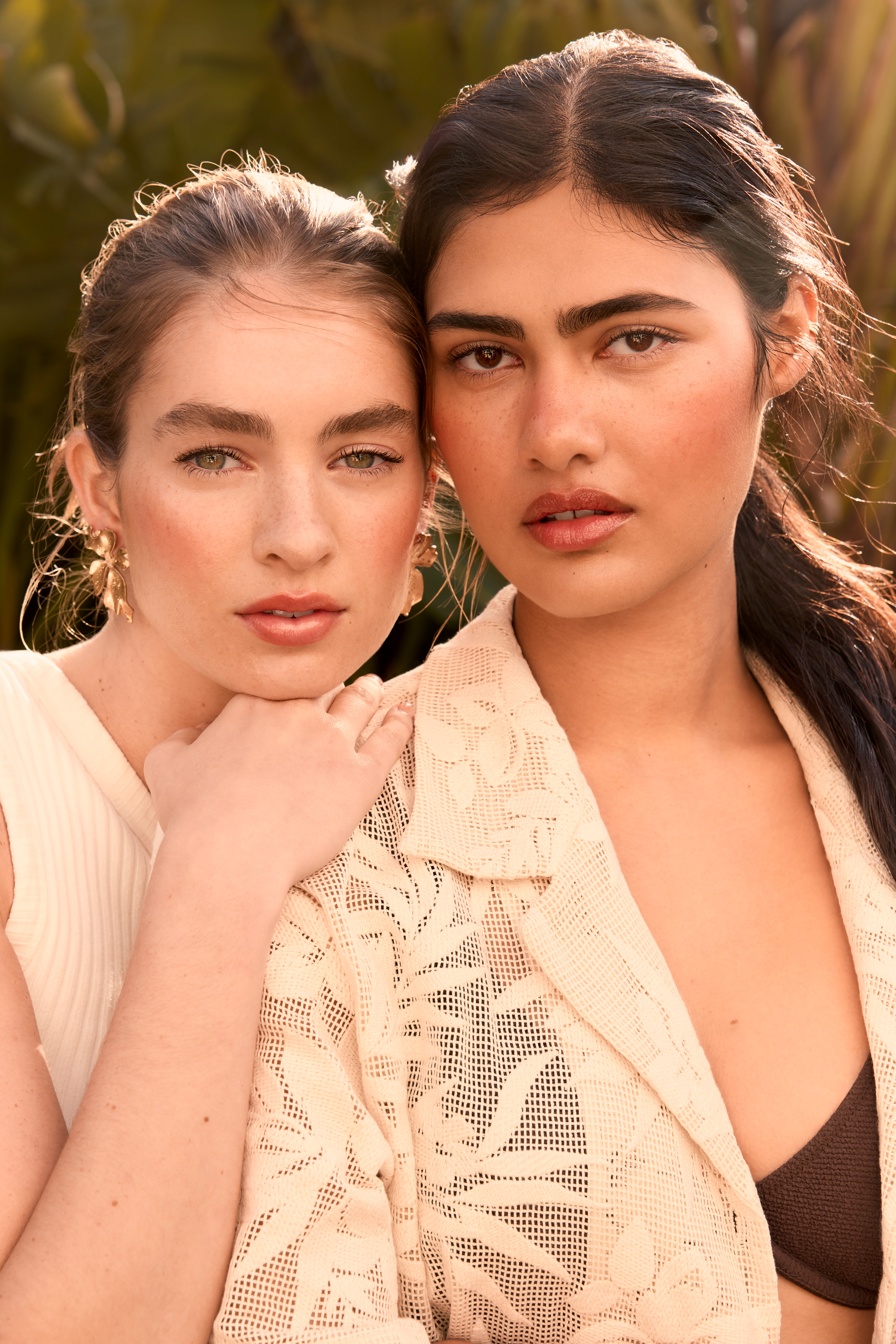 Two women pose outdoors in a close-up shot, one with a cream-colored floral jacket and a dark top, the other in a ribbed tank top.