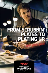 A young male chef in a dark uniform plating food in a commercial kitchen with spherical pendant lights overhead