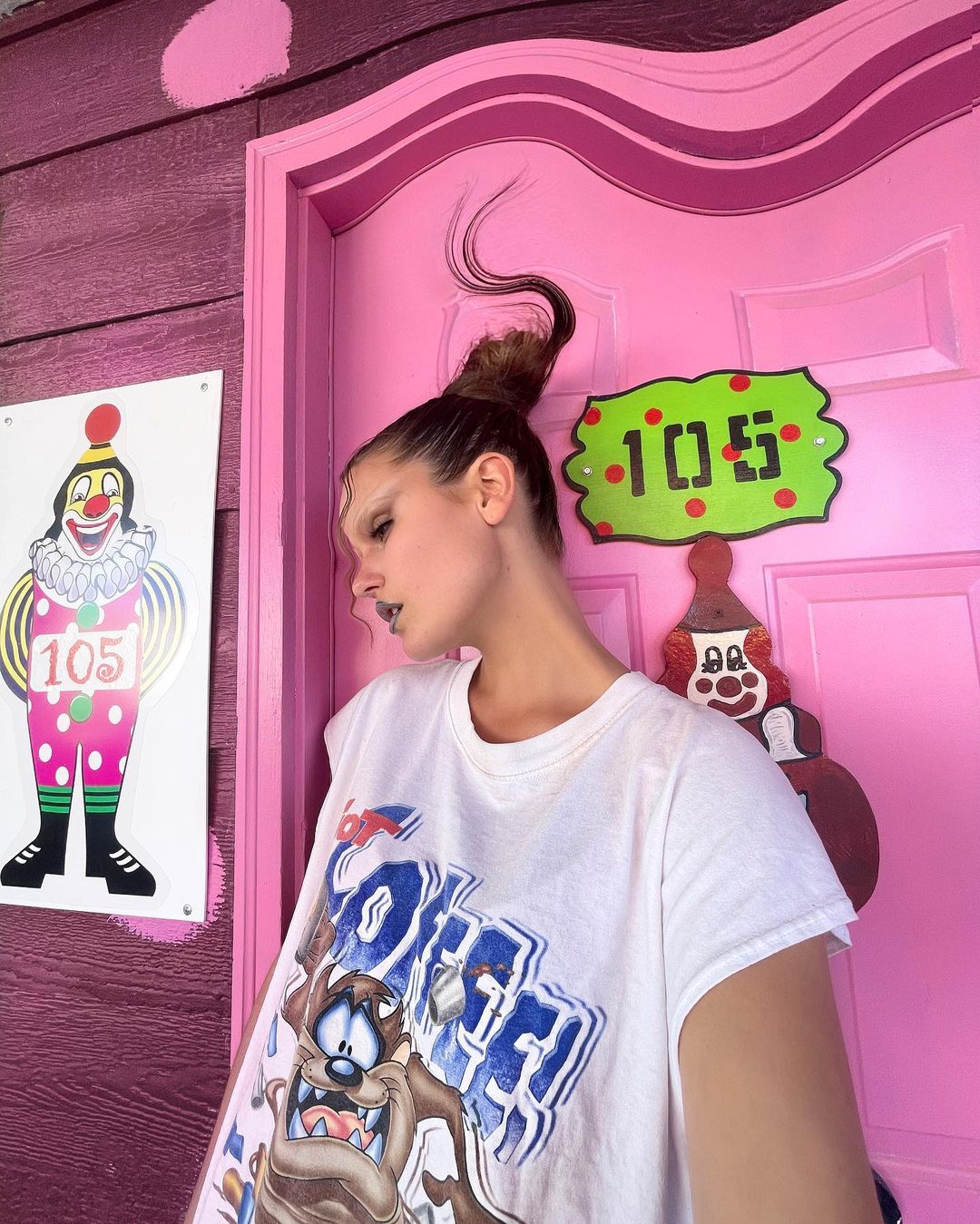Woman with unique hairstyle poses near pink door and clown art, wearing a white graphic tee shirt in an indoor setting.