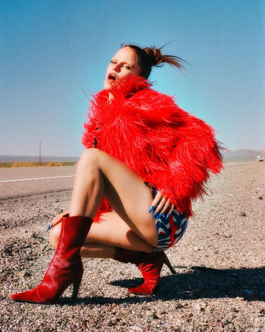 An adult female in a red fuzzy jacket and boots poses outdoors in a crouching position on a road with a light-blue sky backdrop.