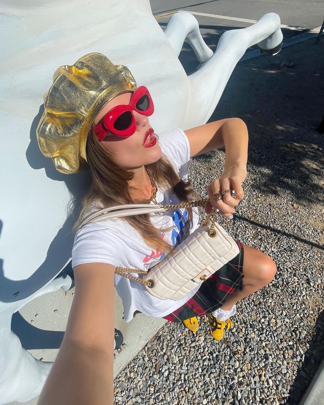 Adult female poses with modern hat and red sunglasses alongside a white horse statue on a sunny day in an outdoor urban environment.