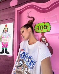 Woman in white graphic t-shirt leaning against a pink door with a whimsical updo hairstyle and clown signage