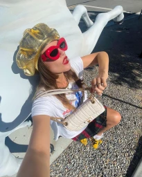 Woman wearing a metallic gold beret and red sunglasses posing near a large white sculpture outdoors.