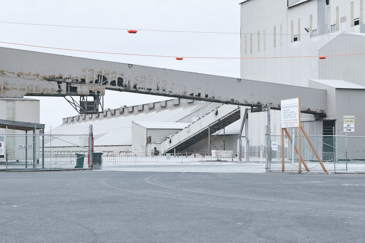 Exterior shot of a large industrial complex with a connecting elevated walkway over a paved lot under an overcast sky.