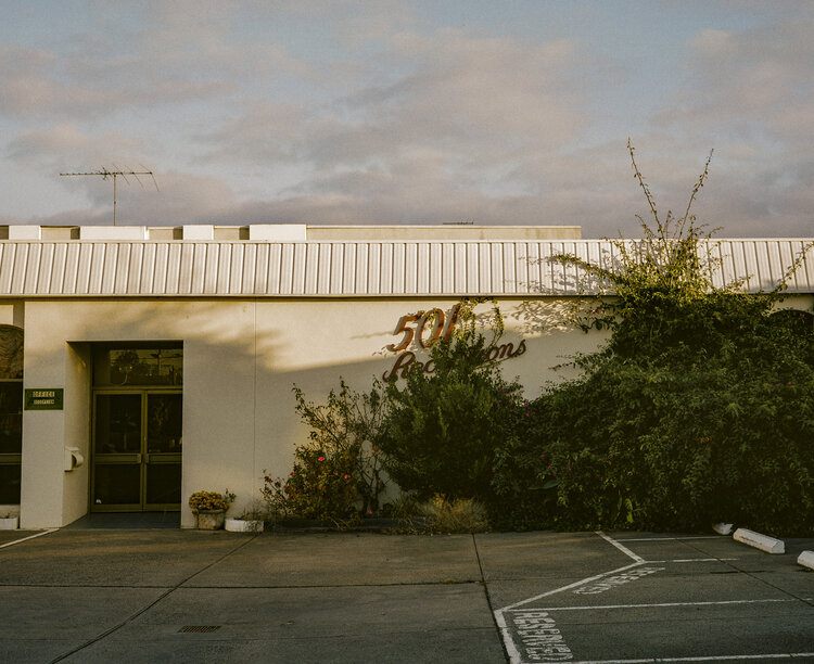 Exterior of a building with a sign reading '501 Rooms,' surrounded by lush greenery and a parking area on an overcast day.