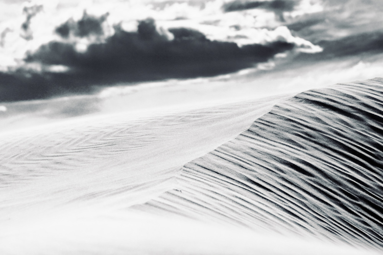 Monochromatic landscape photograph of a rippled sand dune under a cloudy sky, evoking texture and stark natural beauty in black and white.
