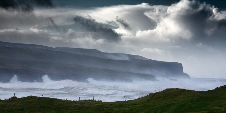 Scenic view of waves crashing against a rocky cliffside under a dramatic, cloudy sky, captured in a wide, landscape-oriented shot.