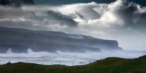 Dramatic coastal landscape with churning ocean waves crashing against dark rocky cliffs beneath a heavy overcast sky