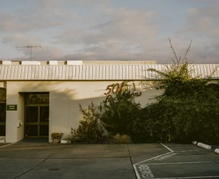 Exterior shot of a white commercial building with lush green bushes and an asphalt parking lot under a cloudy sunset sky.