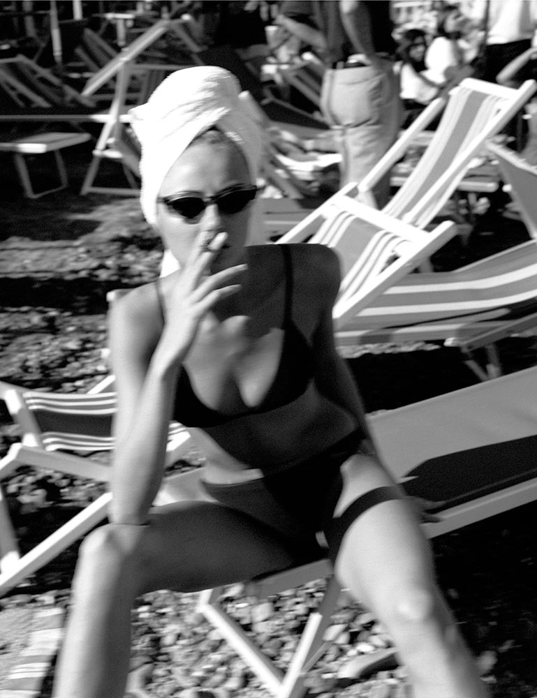 Black and white photo of a woman in a bikini sitting in a beach chair with a towel on her head and smoking a cigarette.