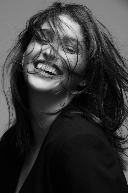 Black and white portrait of a smiling woman with windblown hair, wearing a black jacket against a grey background.