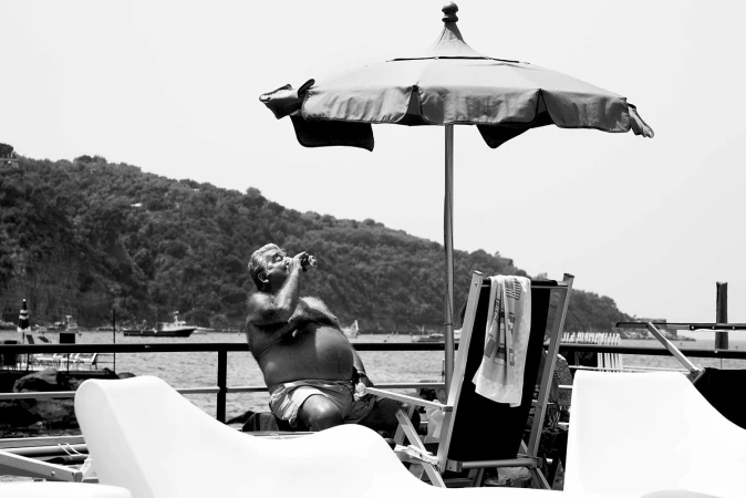 Monochrome image of an older man relaxing in a beach chair drinking from a bottle under an umbrella by the water.