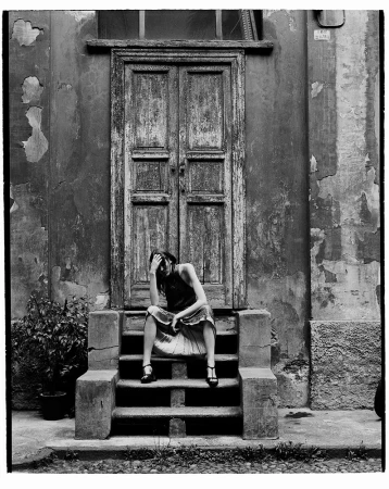 Black and white photo of a woman in dress sitting on concrete steps leading to a weathered door, with peeling plaster walls.